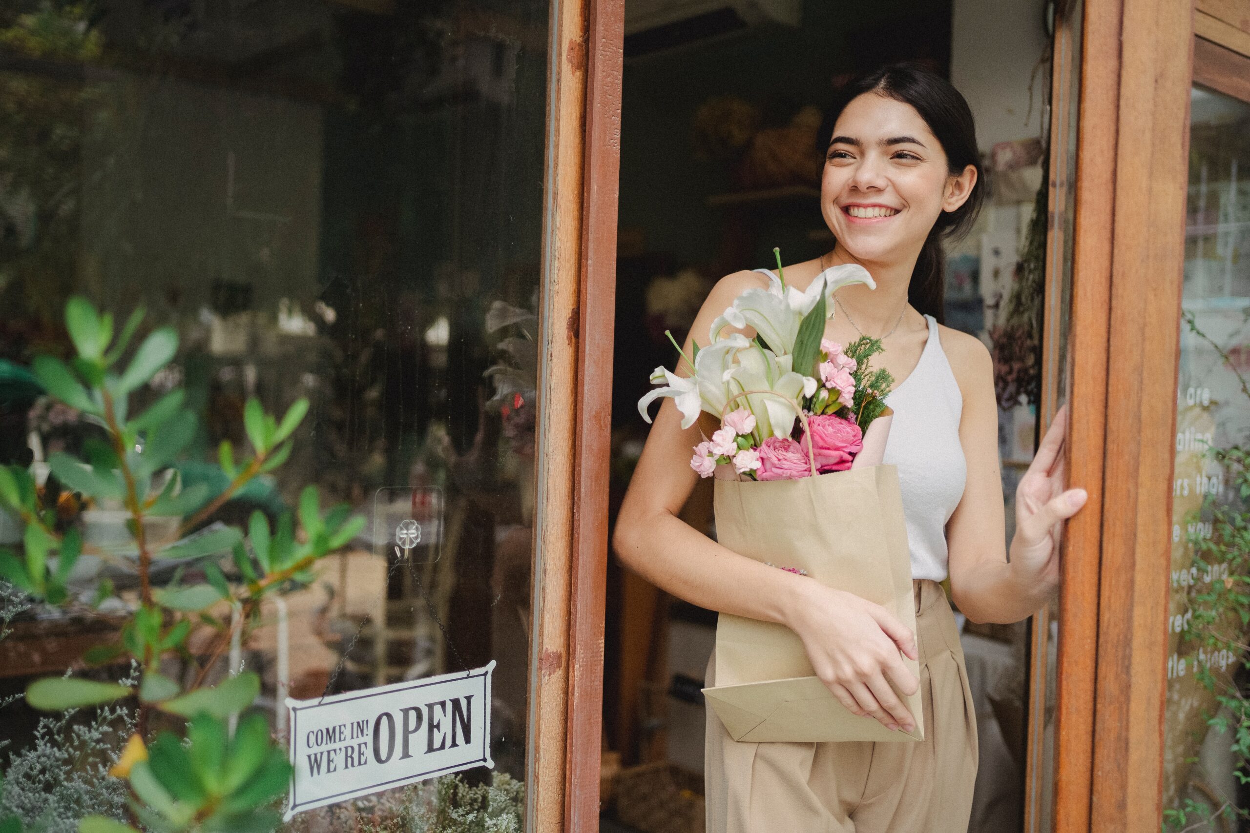 woman standing at the door of a store with an Open sign, smiling and holding a bag of flowers